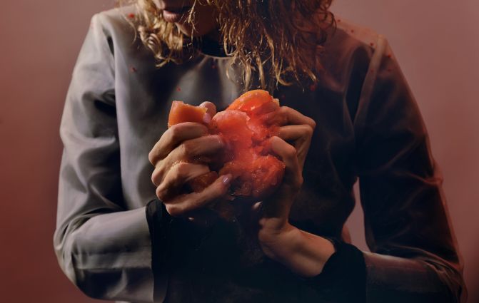 Eine Frau schaut zur Seite, man sieht ihren Körper nur vom Mund abwärts. Sie hat lange braunen Haaren. In den Händen hat sie eine rote Tomate, die sie in den Händen vermantscht. Es rinnt an den Händen die Tomatenflüssigkeit runter.