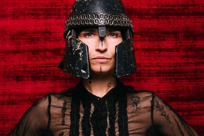 Michèle Rohrbach mit ernstem Blick trägt einen mittelalterlichen schwarzen Helm mit Metallverzierungen. Sie steht vor einem roten, samtartigen Hintergrund und trägt eine transparente schwarze Bluse.