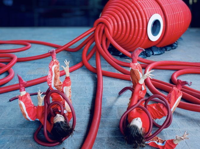 Zwei rot angezogene Spielerinnen liegen auf dem Rücken auf einem Steinboden zwischen roten Plastiktentakel von einem roten, großen Plastiktintenfisch, der daneben liegt.