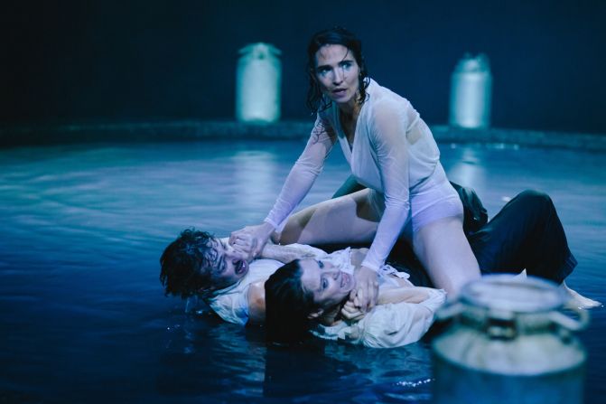 Drei Personen befinden sich in einem flachen Wasserbecken. Michèle Rohrbach in weisser Kleidung sitzt auf den anderen beiden und hält sie fest. Im Hintergrund stehen Milchkannen.