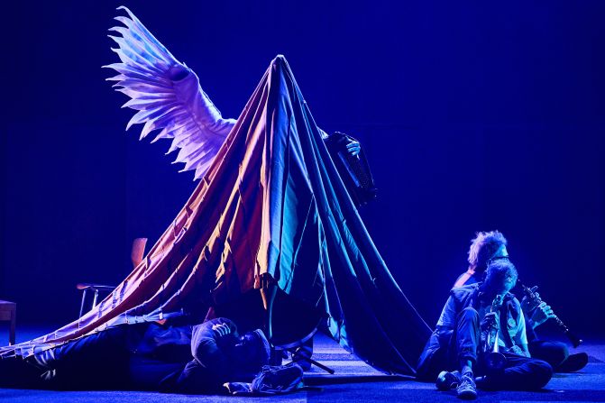 Eine Bühne in blau-violettem Licht zeigt eine Szene mit mehreren Personen, die am Boden liegen oder sitzen. Im Zentrum erhebt sich eine Gestalt unter einem grossen Tuch mit weissen Engelsflügeln.