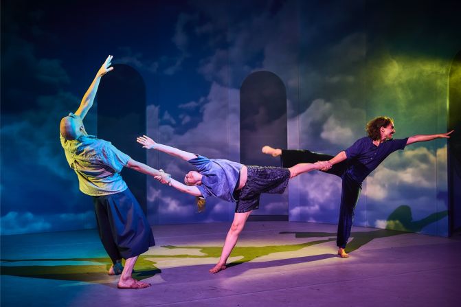 Drei zeitgenössische Tänzer:innen inmitten einer Performance, balancierend und einander haltend in einer dynamischen Pose vor einem traumhaften Hintergrund aus Wolken und Bögen.