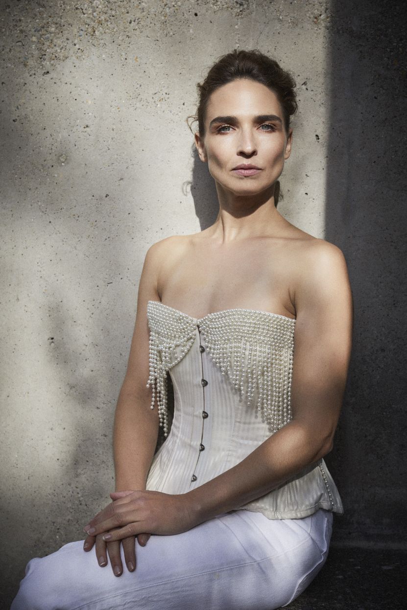 Michèle Rohrbach sitzt vor einer Betonwand im Licht-Schatten-Spiel. Sie trägt ein elegantes, schulterfreies Korsett mit Perlendekor und eine weisse Hose. Die Beine sind übereinander geschlagen und ihre Hände ruhen auf dem Oberschenkel.