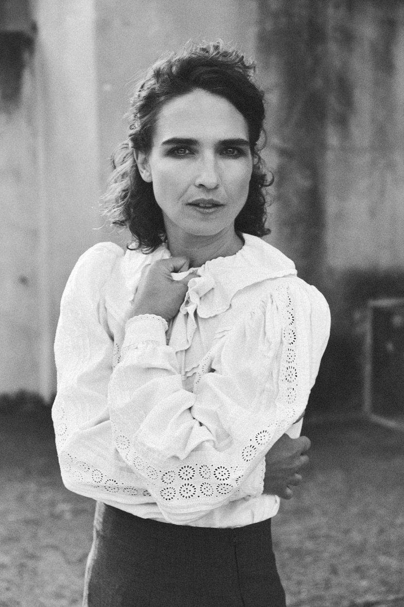 Michèle Rohrbach in weisser Bluse mit Lochstickerei steht mit verschränkten Armen und nachdenklichem Blick vor einer verwitterten Wand.