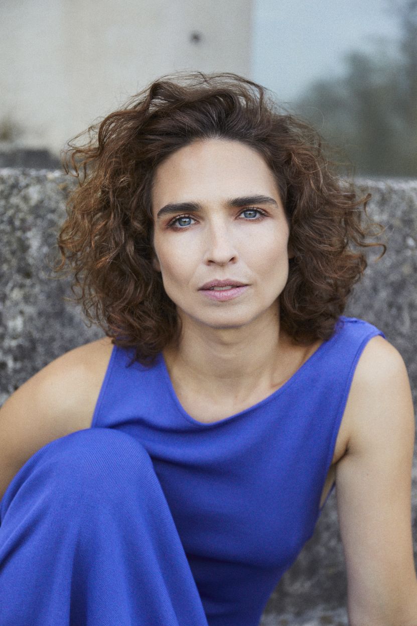 Michèle Rohrbach mit lockigem braunem Haar und intensivem Blick sitzt vor einer grauen Steinmauer. Sie trägt ein ärmelloses, leuchtend blaues Kleid und schaut direkt in die Kamera.