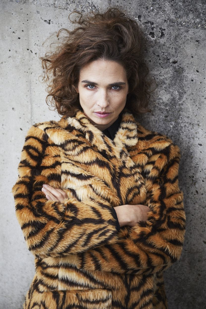 Michèle Rohrbach mit lockigem Haar steht mit verschränkten Armen vor einer grauen Betonwand. Sie trägt einen auffälligen Kunstpelzmantel im Tigermuster und blickt selbstbewusst in die Kamera.